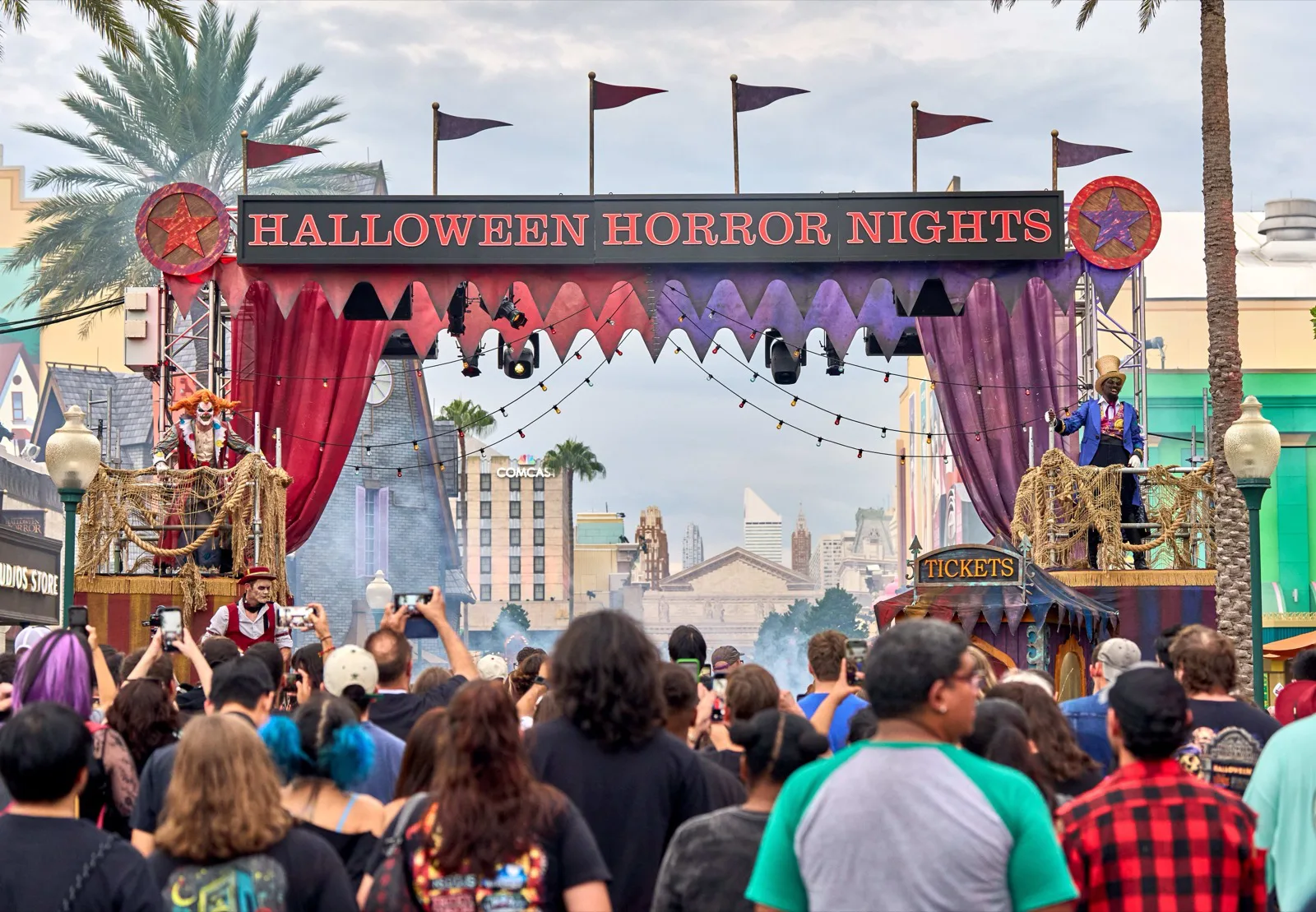 Halloween Horror Nightsの装飾ゲート前に集まる来場者と、両脇に立つカーニバル風の出演者